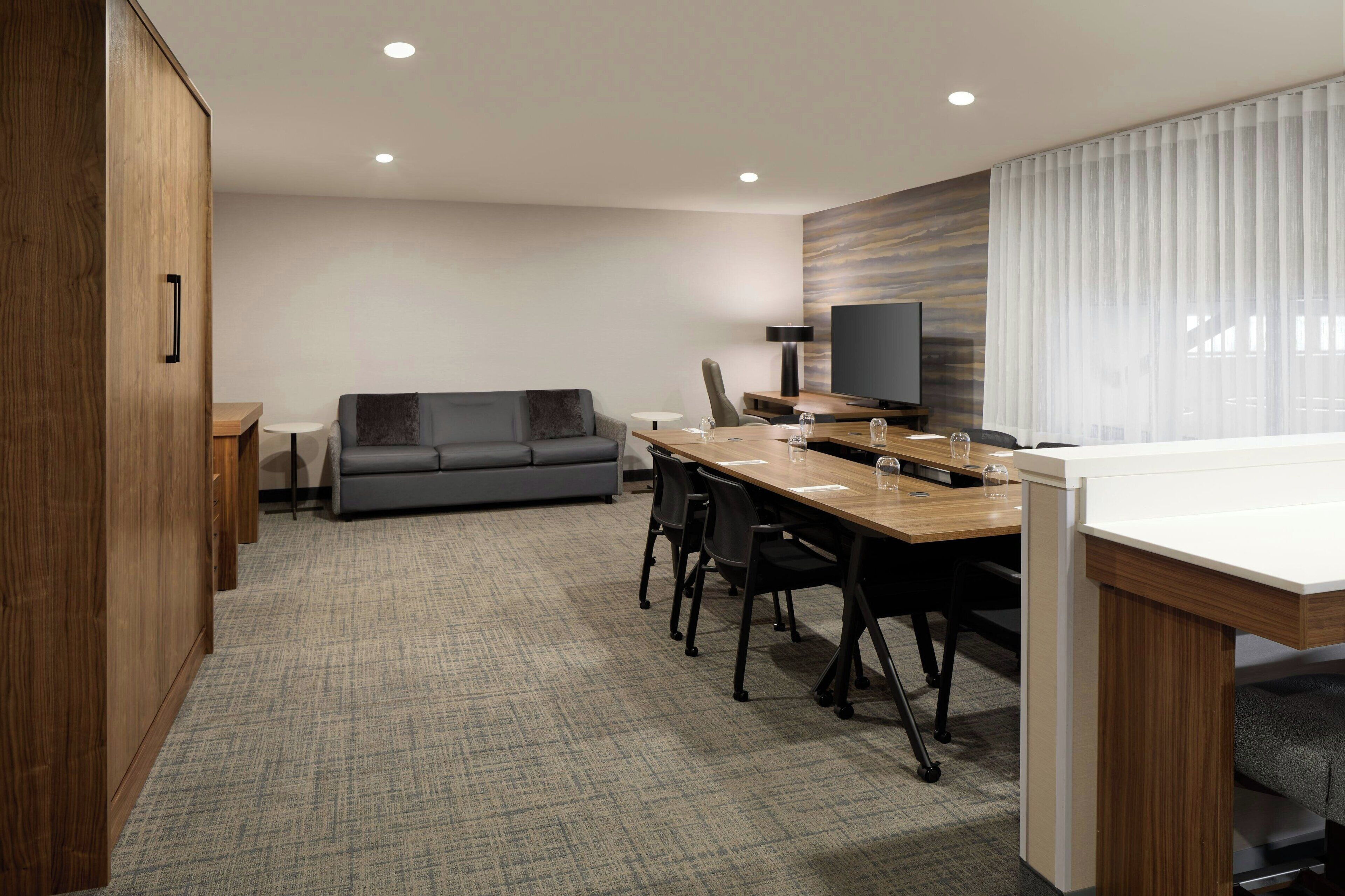 Een moderne hotelkamer met een houten garderobe, een grijze bank, een eettafel met stoelen, een flatscreen televisie en een groot raam met gordijnen.