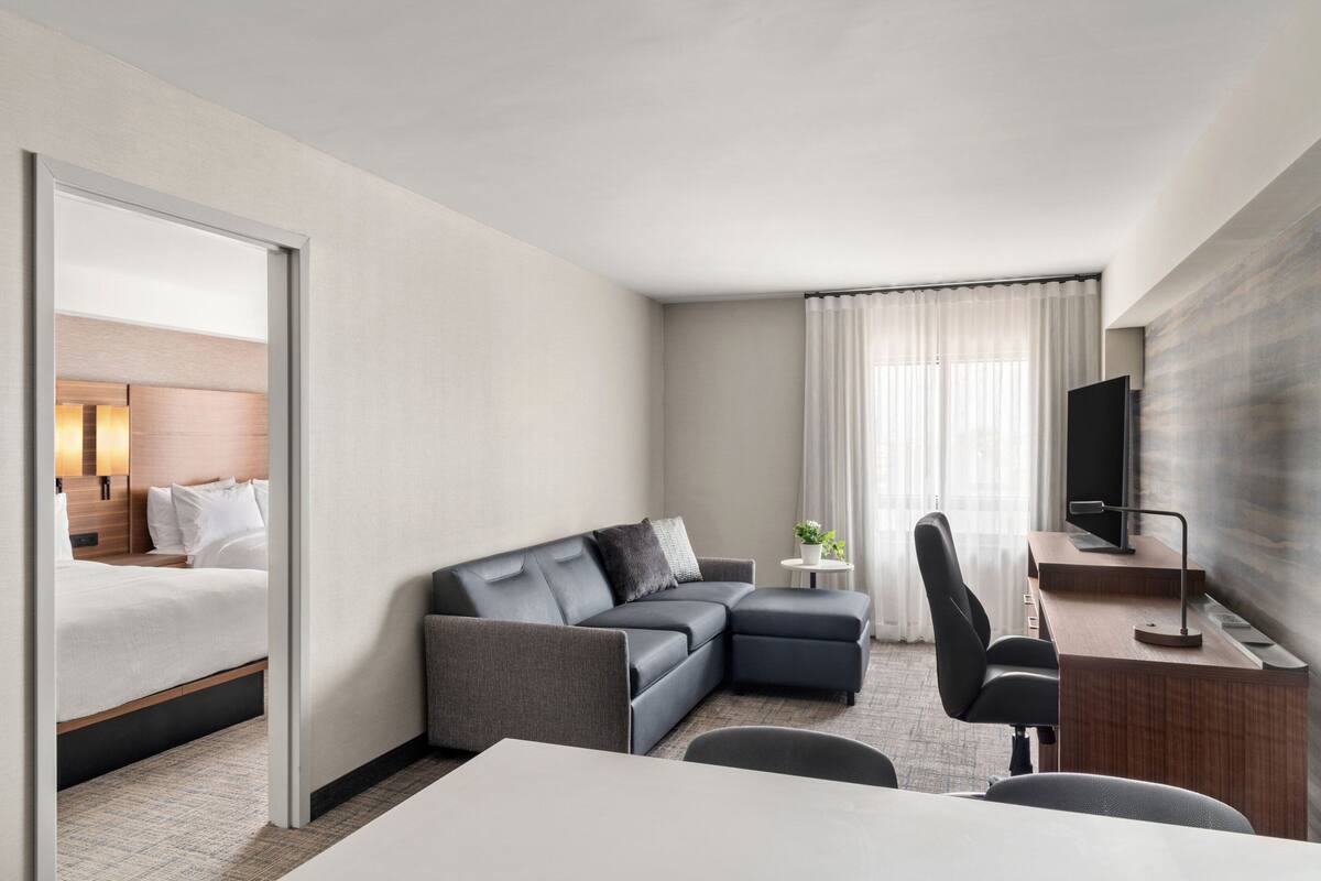 A modern hotel room with a bed, gray sofa, desk, and a TV.