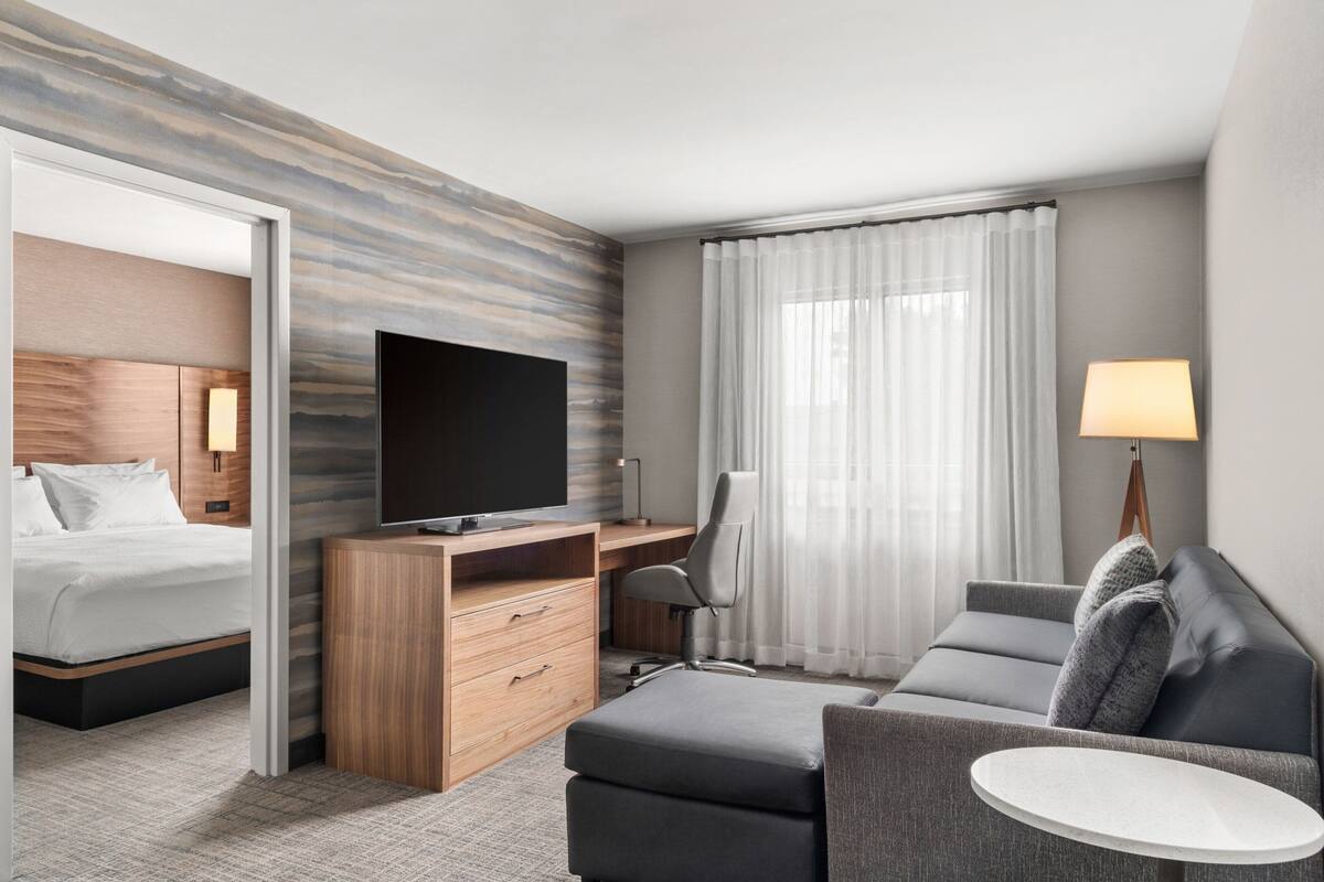 A modern hotel room with a bed, a desk with a TV, a sofa, a small table, and a lamp.