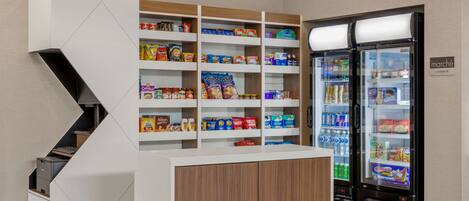 A kitchenette area with a white and wooden counter, shelves stocked with snacks and beverages, and two black beverage dispensers.
