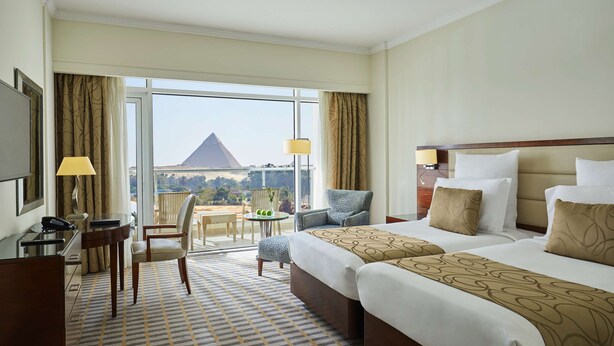 A hotel room with two beds, a desk, a chair, a small table, and a view of a mountain. - Cairo Pyramids Hotel (Giza)