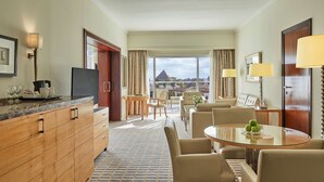 A hotel room with a dining area, a TV, a wooden cabinet, and a view of a pyramid. - Cairo Pyramids Hotel (Giza)