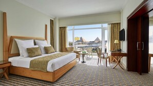 A hotel room with a large bed, a desk with a chair, a small table with a lamp, and a view of pyramids through the window. - Cairo Pyramids Hotel (Giza)