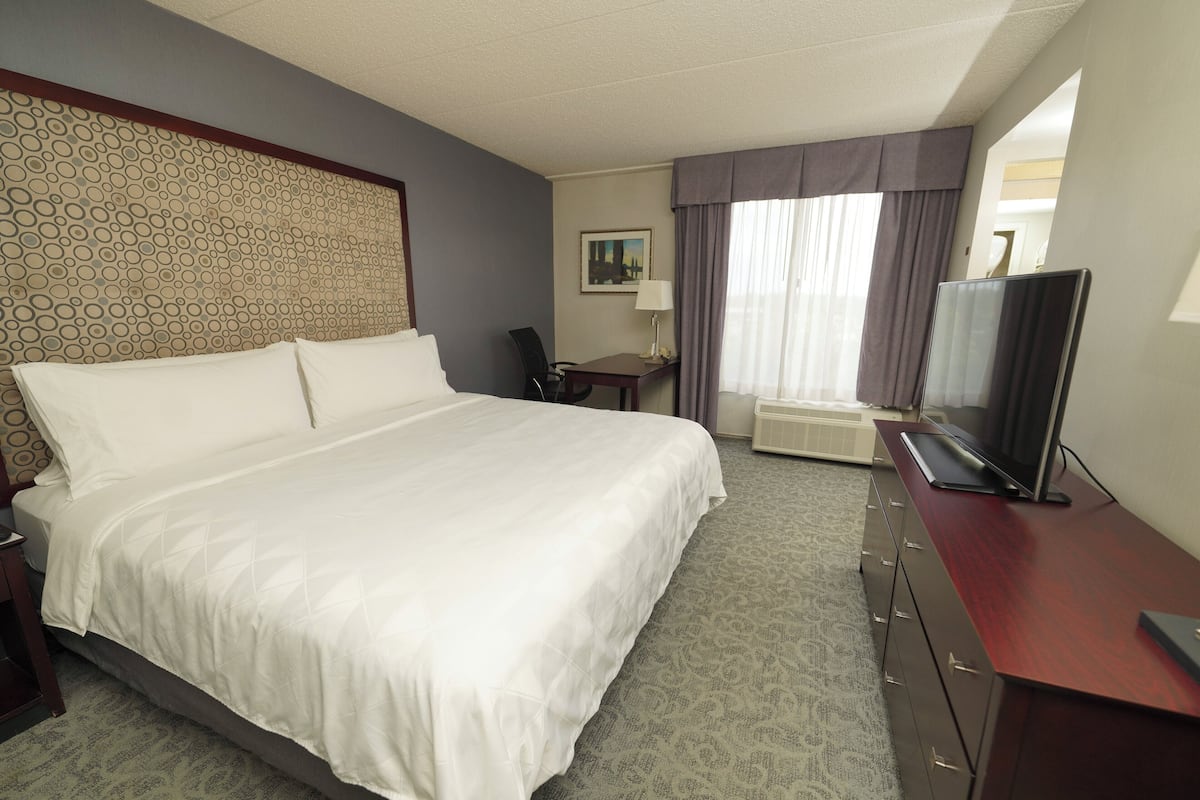 A hotel room with a large bed, a desk with a flat-screen TV, a chair, a lamp, and a painting on the wall.