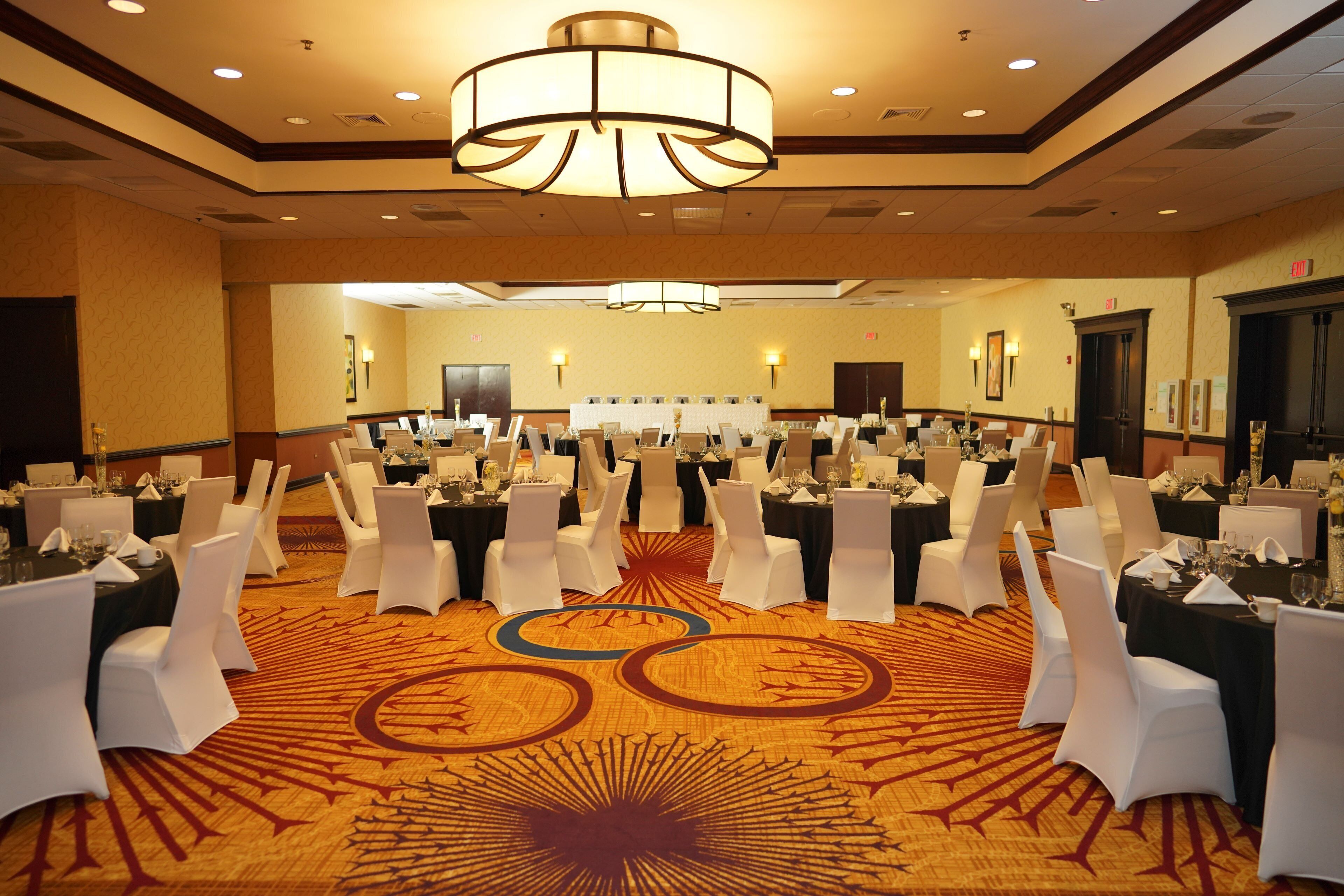 A banquet hall with round tables, white chairs, and a patterned carpet.
