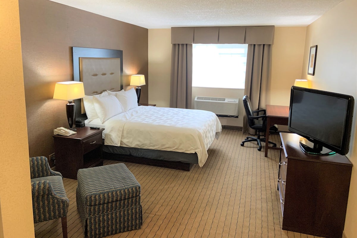 A hotel room with a bed, two bedside lamps, a desk with a computer, a chair, an ottoman, and a TV.