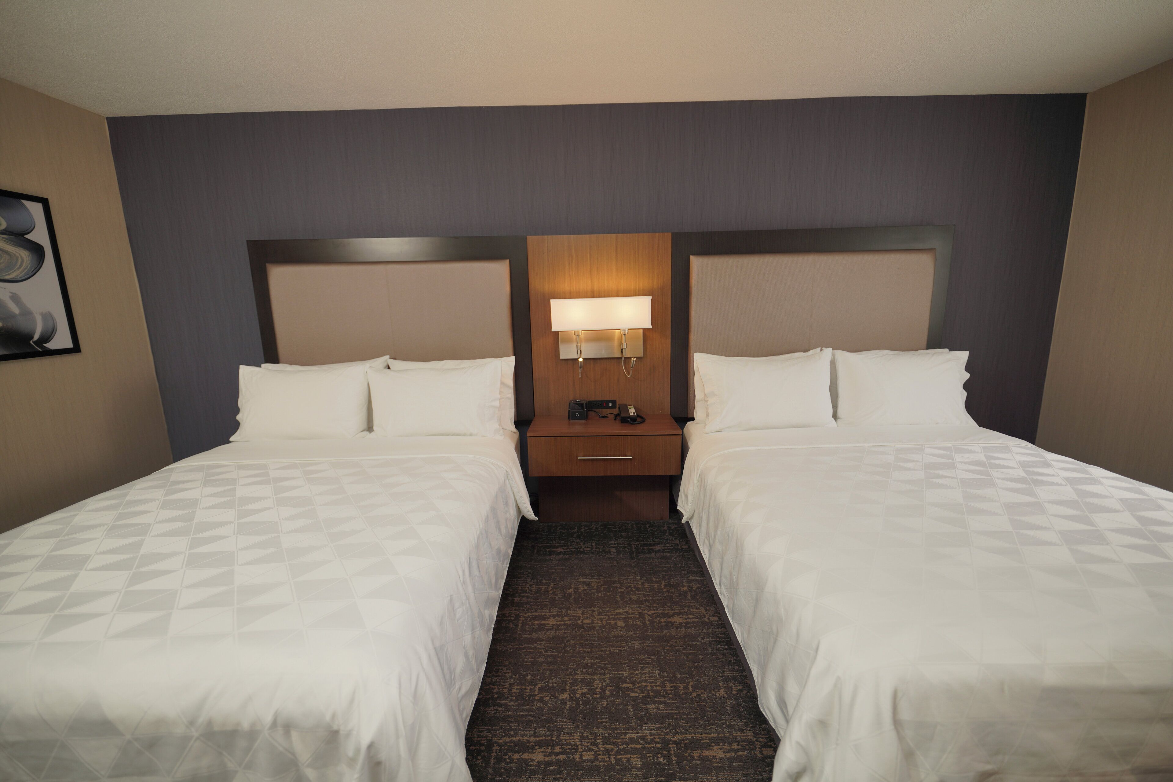 A hotel room with two beds, a nightstand, a lamp, and a framed picture on the wall.
