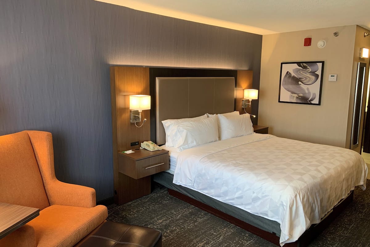 A hotel room with a bed, two bedside lamps, a nightstand, a telephone, a chair, and a wall-mounted artwork.