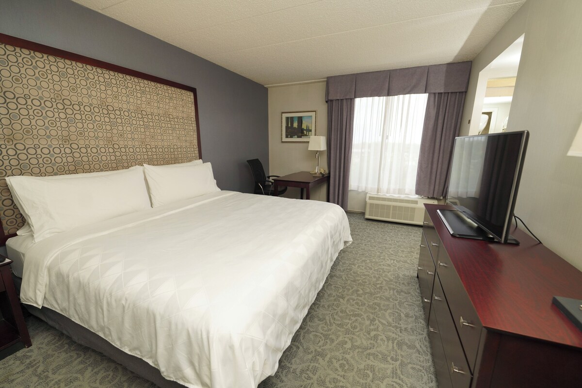 Een hotelkamer met een groot bed, een bureau met een flatscreen televisie, een stoel, een lamp en een schilderij aan de muur.