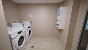 A laundry room with two front-loading washing machines, a dryer, and a wall-mounted soap dispenser. - Holiday Inn & Suites Chicago-Carol Stream (Wheaton) by IHG (Carol Stream)