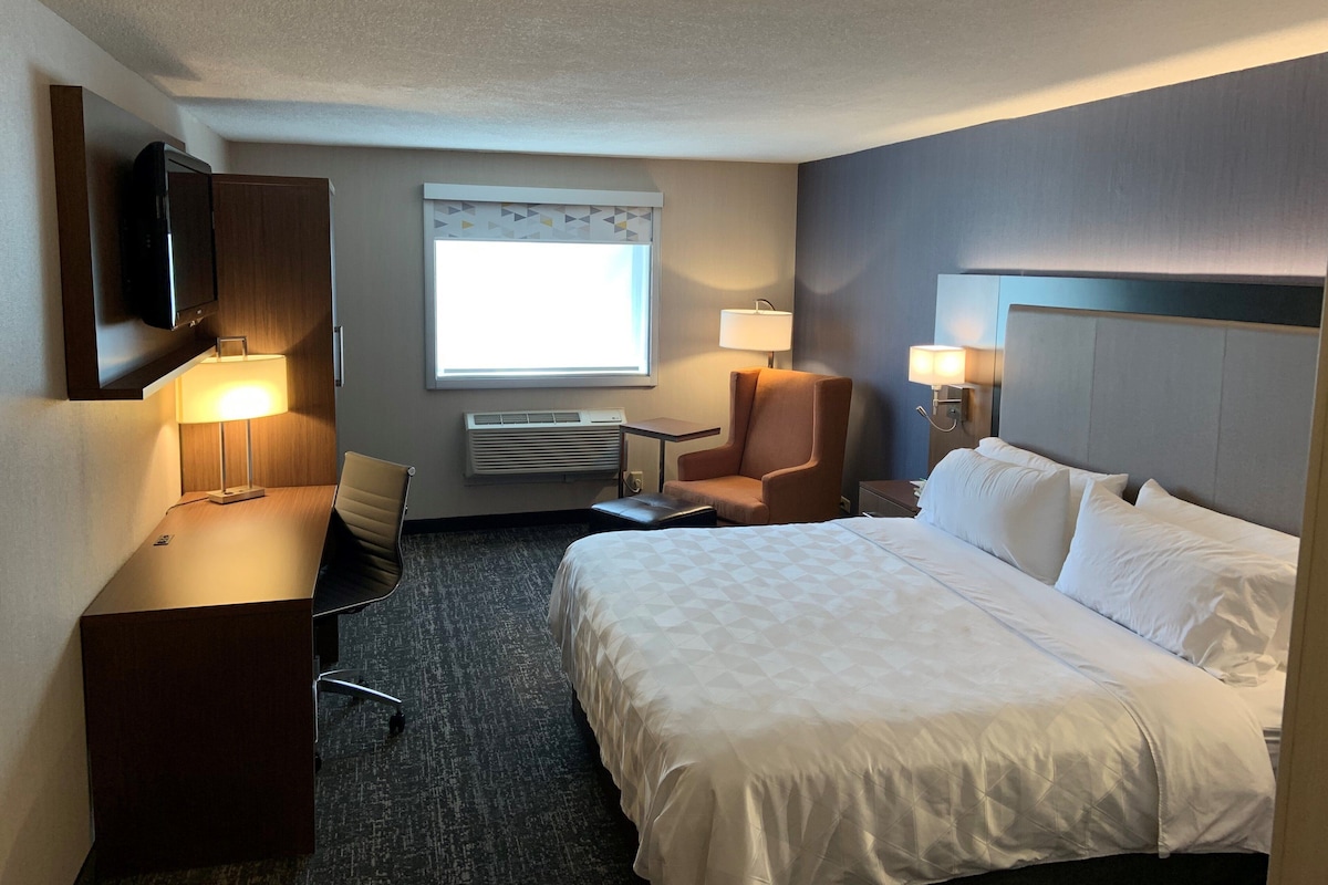 Een hotelkamer met een bed, een bureau met stoel, een televisie, een raam met jaloezieën en een wandlamp.