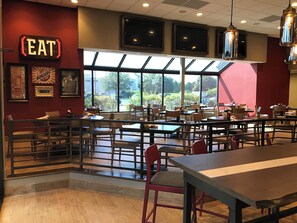 A restaurant interior with tables and chairs, a large window, and a sign that says 'EAT'. - Holiday Inn & Suites Chicago-Carol Stream (Wheaton) by IHG (Carol Stream)
