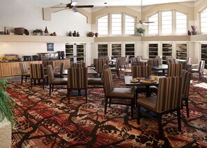 A spacious dining area with striped chairs and tables, a kitchen in the background, and large windows allowing natural light.
