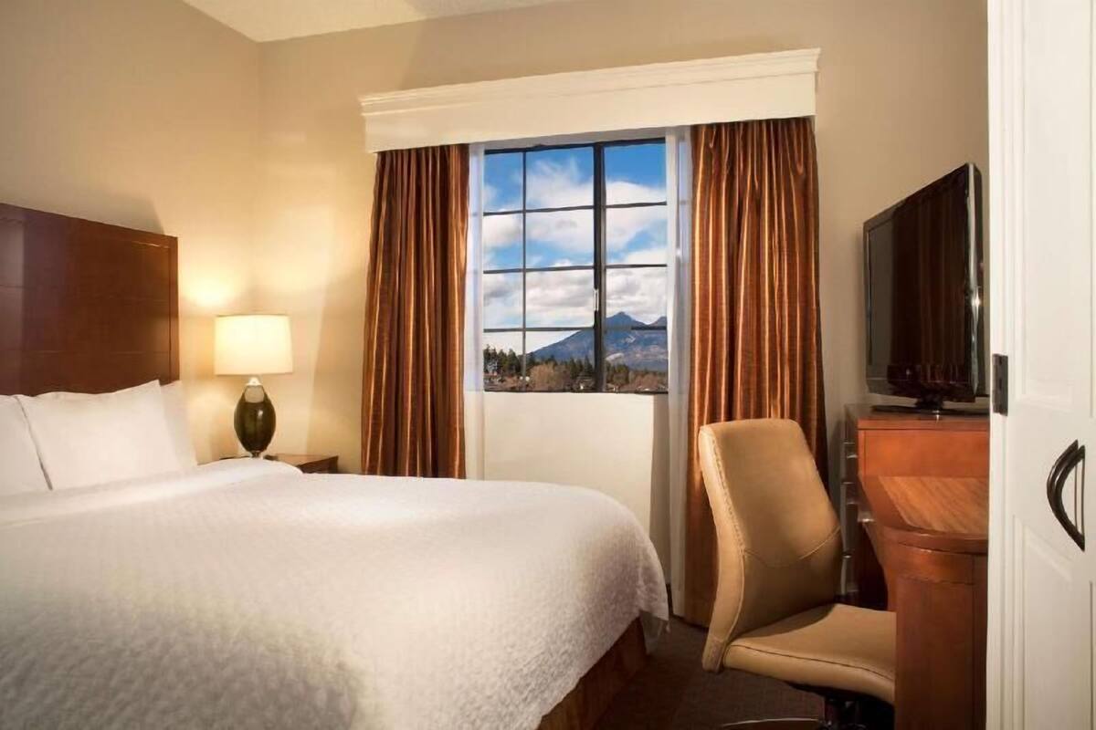 Ein Hotelzimmer mit Bett, Stuhl, Fernseher, Lampe und einem Fenster mit Bergblick.