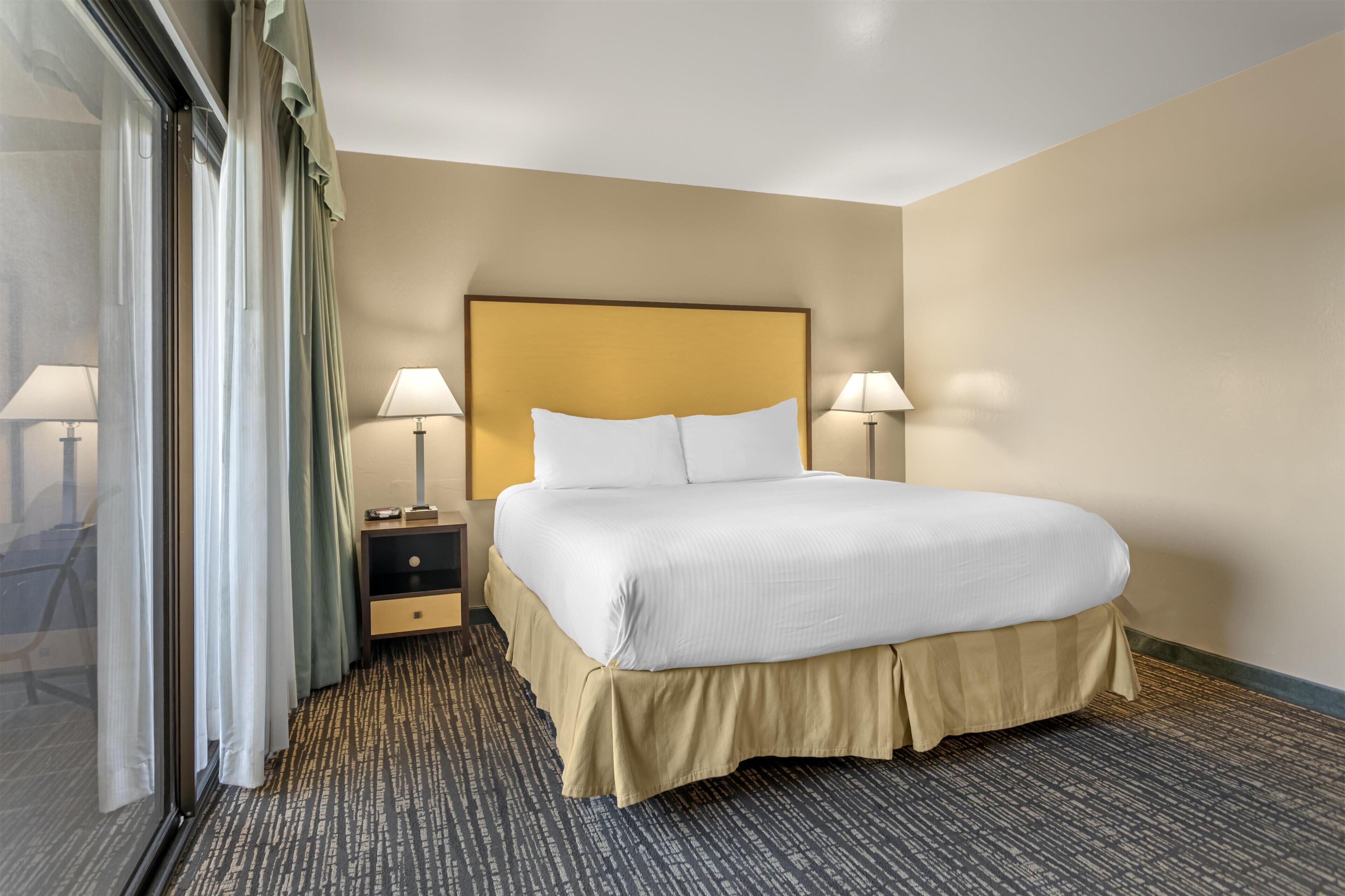Quarto de hotel com uma cama grande, duas luminárias de cabeceira, uma mesinha de cabeceira e vista através de uma porta de vidro.