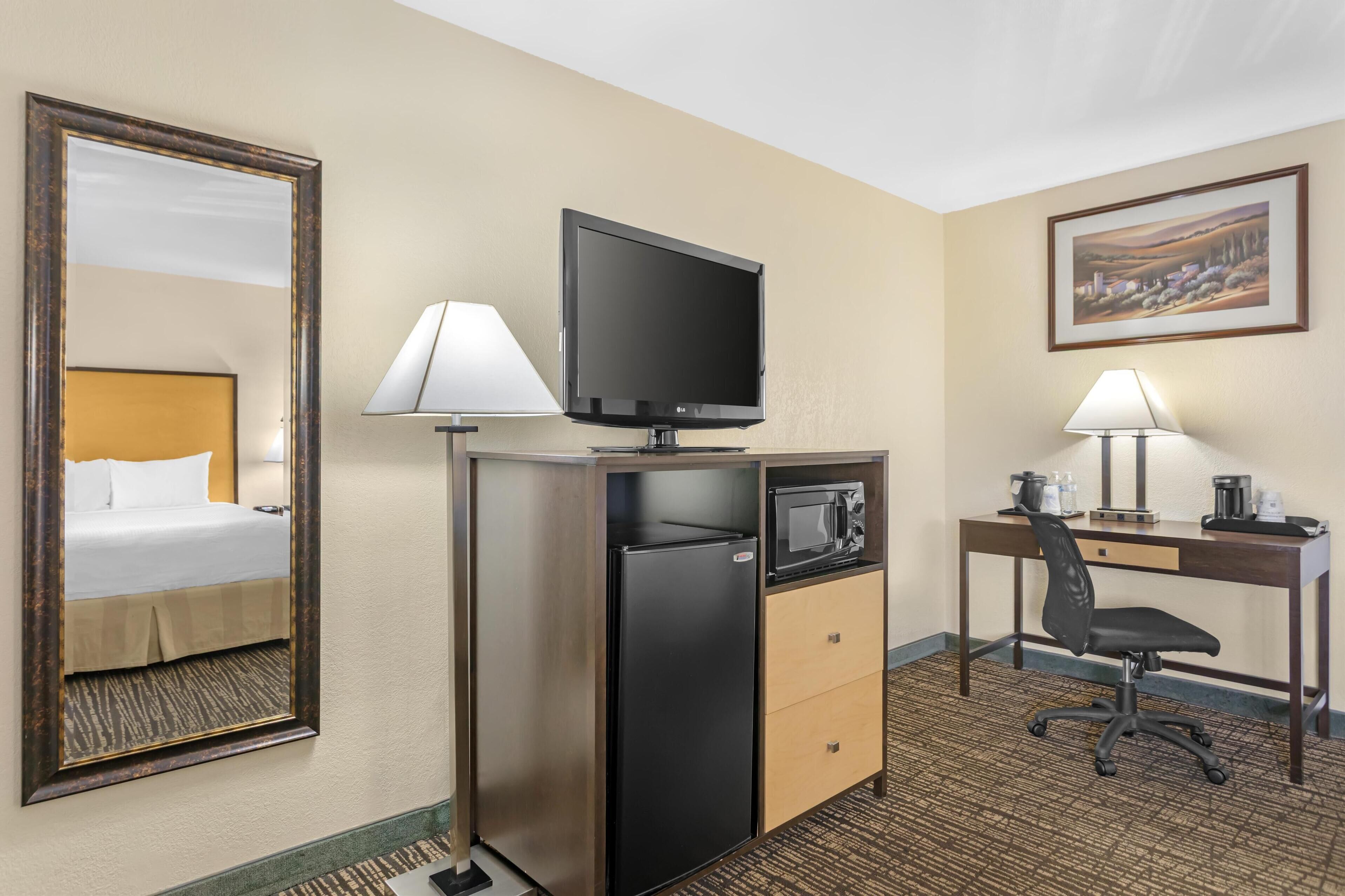 Quarto de hotel com cama, escrivaninha com computador, televisão, frigorífico e micro-ondas.