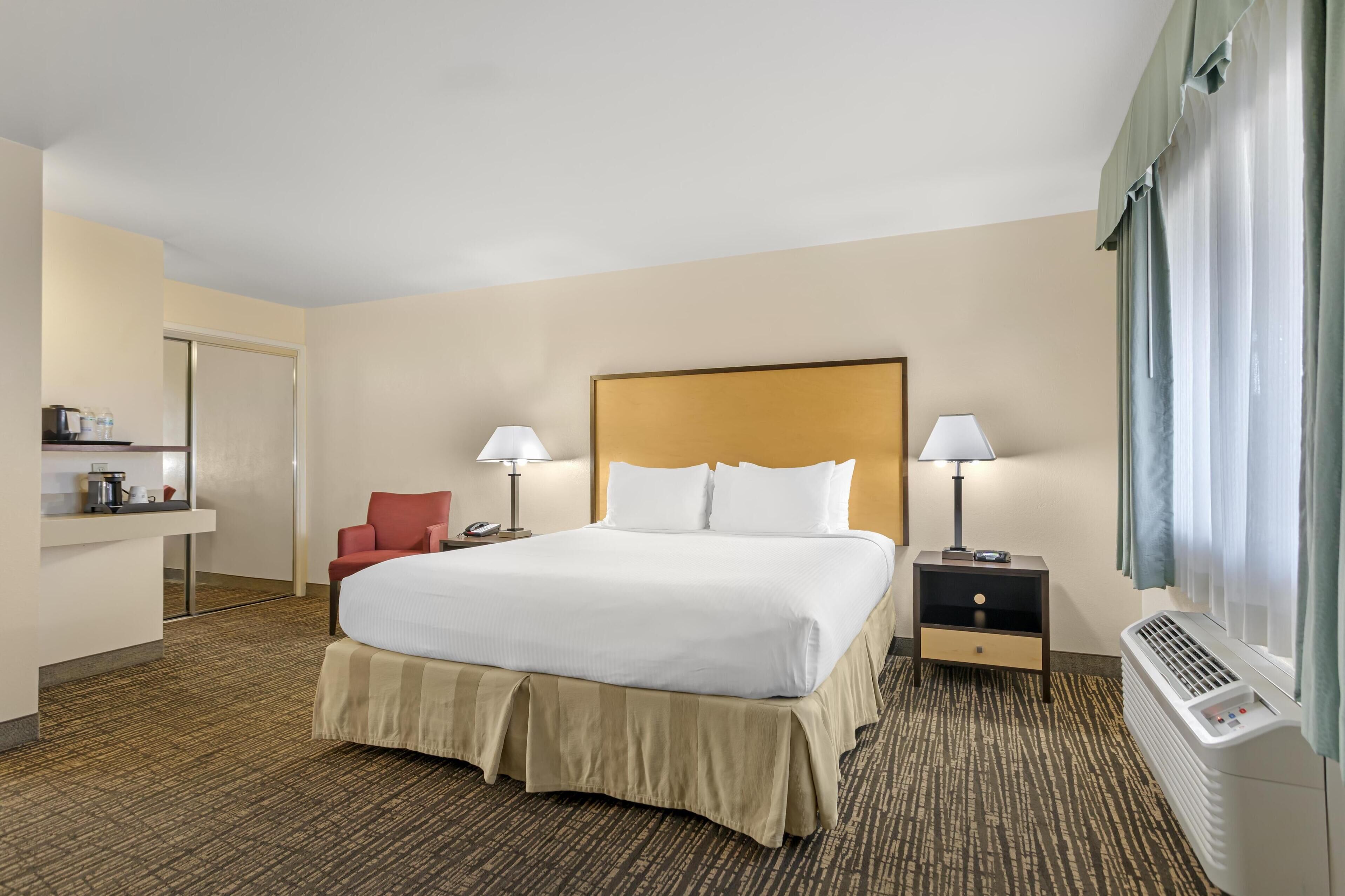 A hotel room with a large bed, two bedside tables with lamps, a red chair, a small desk with a phone, and a window with curtains.