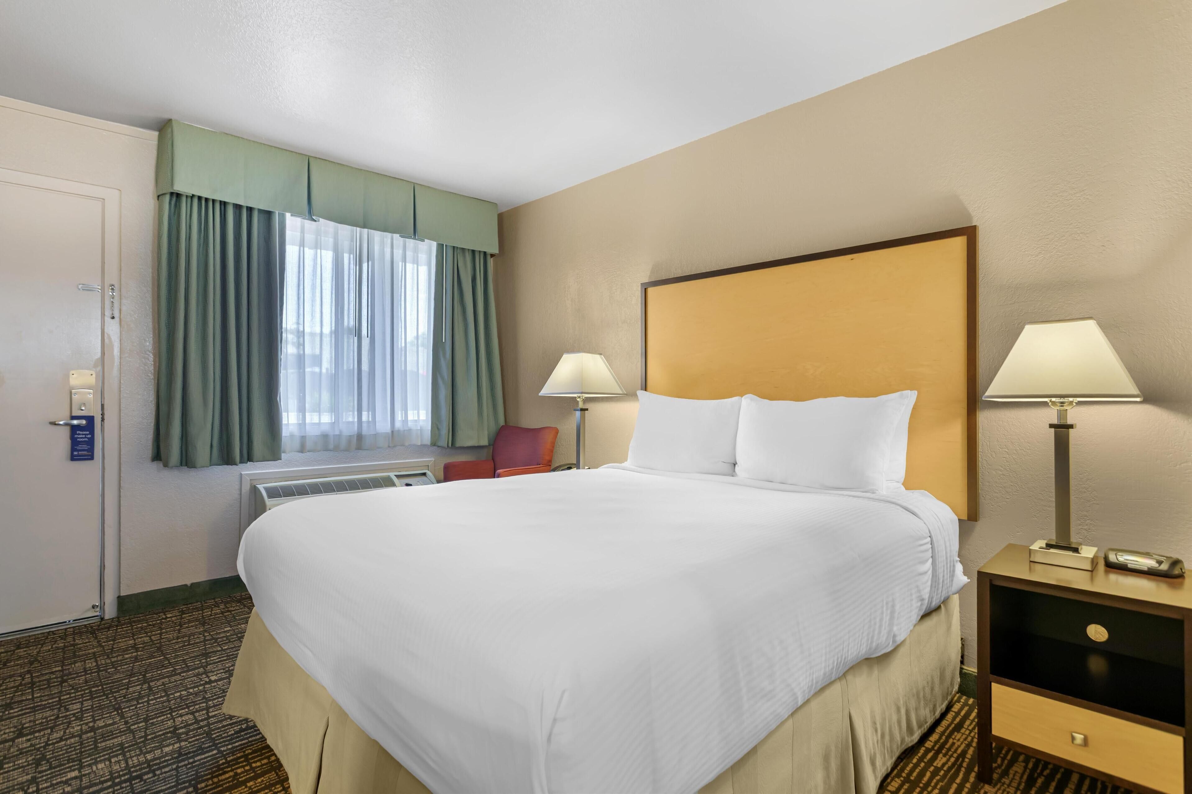 Quarto de hotel com uma cama grande, duas luminárias de cabeceira, uma cadeira vermelha e uma janela com cortinas verdes.