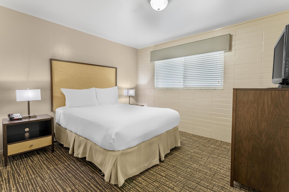 A hotel room with a large bed, two bedside lamps, a wooden nightstand, a television on a wooden cabinet, and a window with blinds.