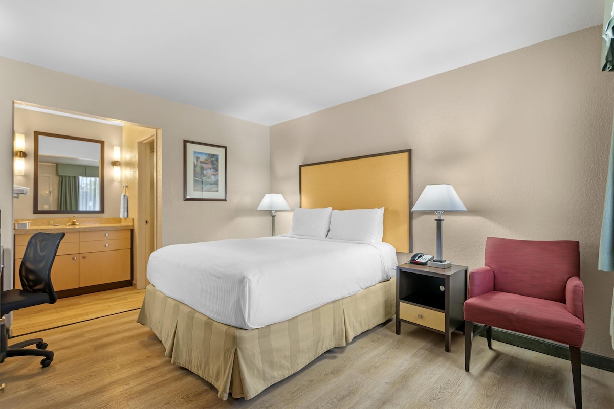 A hotel room with a large bed, a desk with a chair, a nightstand, and a red armchair.