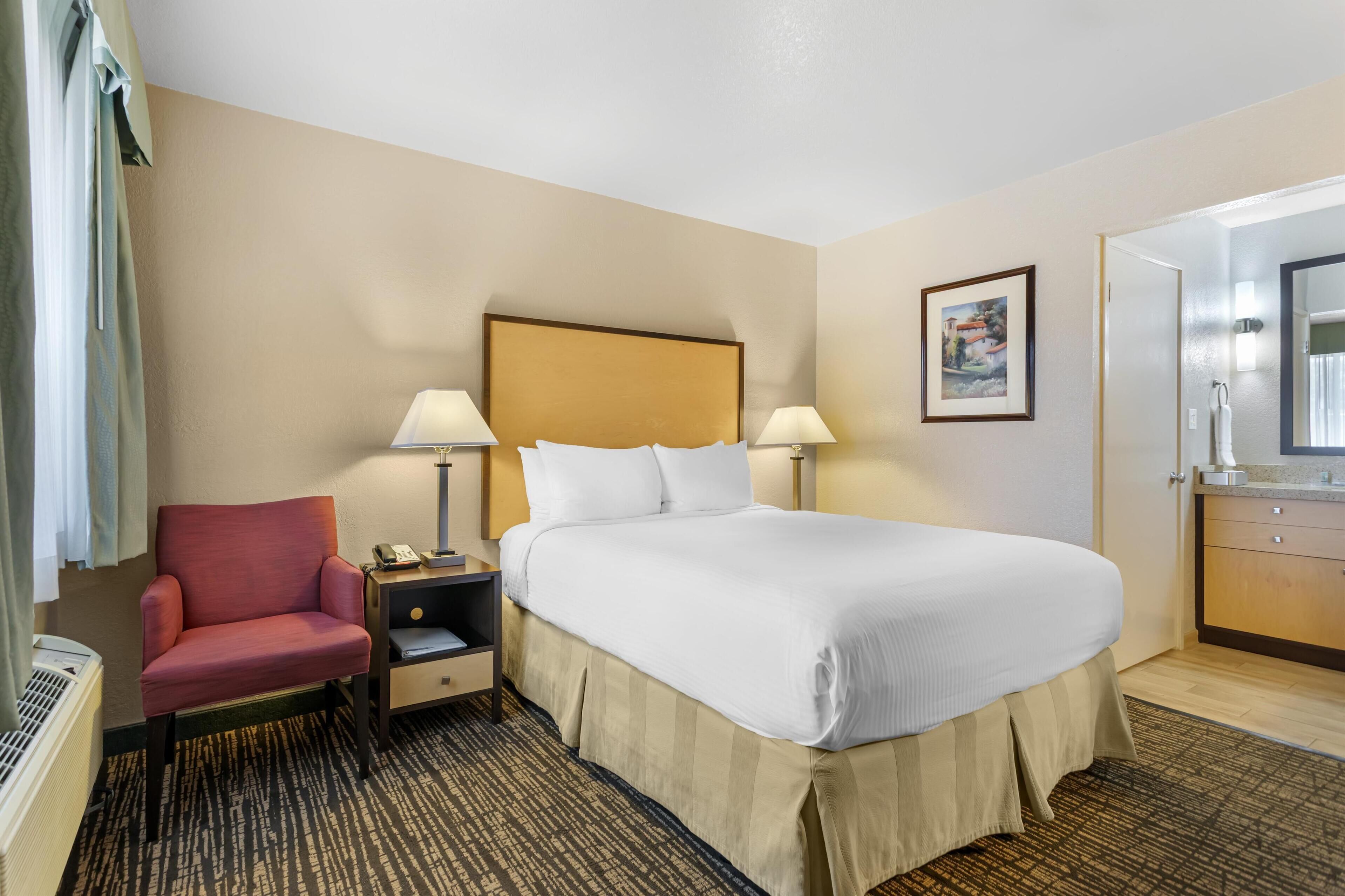 Habitación de hotel con cama, una silla, mesita de noche, lámpara, un cuadro en la pared y un baño visible a través de una puerta abierta.