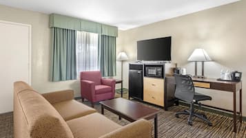 A hotel room with a sofa, a pink armchair, a desk with a computer, a flat-screen TV, a mini-fridge, and a coffee maker.