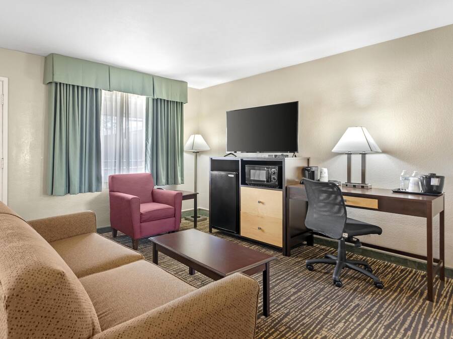 Ein Hotelzimmer mit einer Couch, einem pinken Sessel, einem Schreibtisch mit Computer, einem Flachbildfernseher, einem Minikühlschrank und einer Kaffeemaschine.