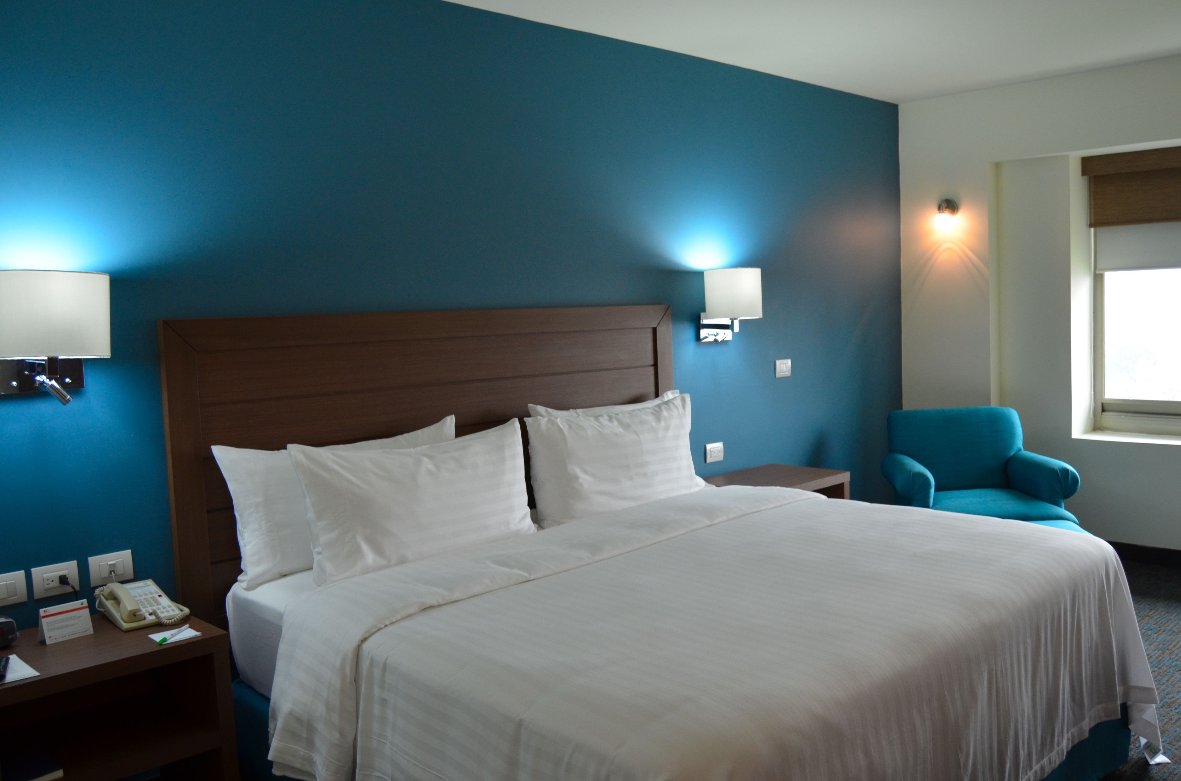 A hotel room with a large bed, a nightstand with a phone, a blue armchair, and wall-mounted lights.
