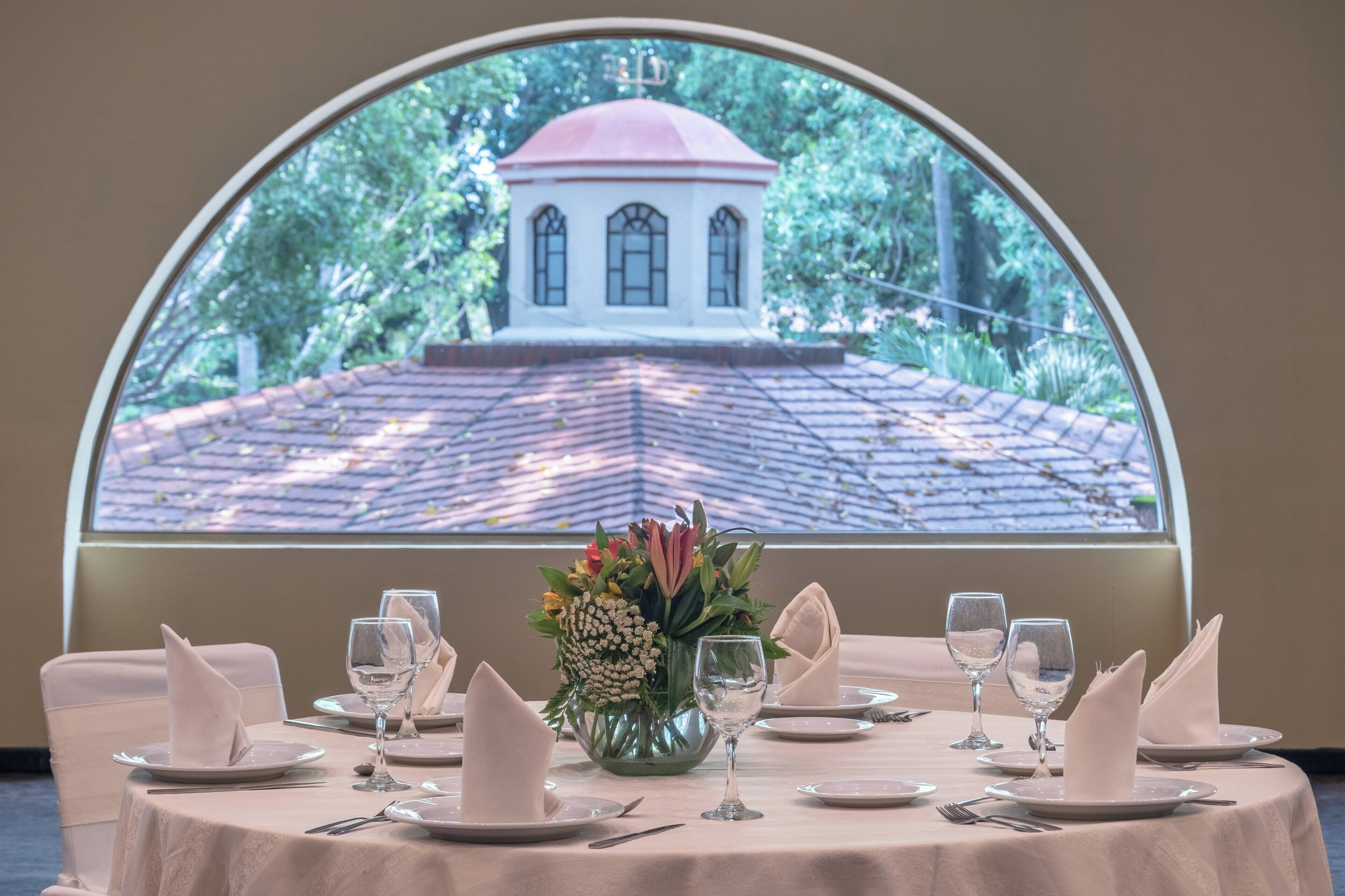Mesa de comedor lista para una comida, con vista a un edificio a través de un amplio ventanal en arco.