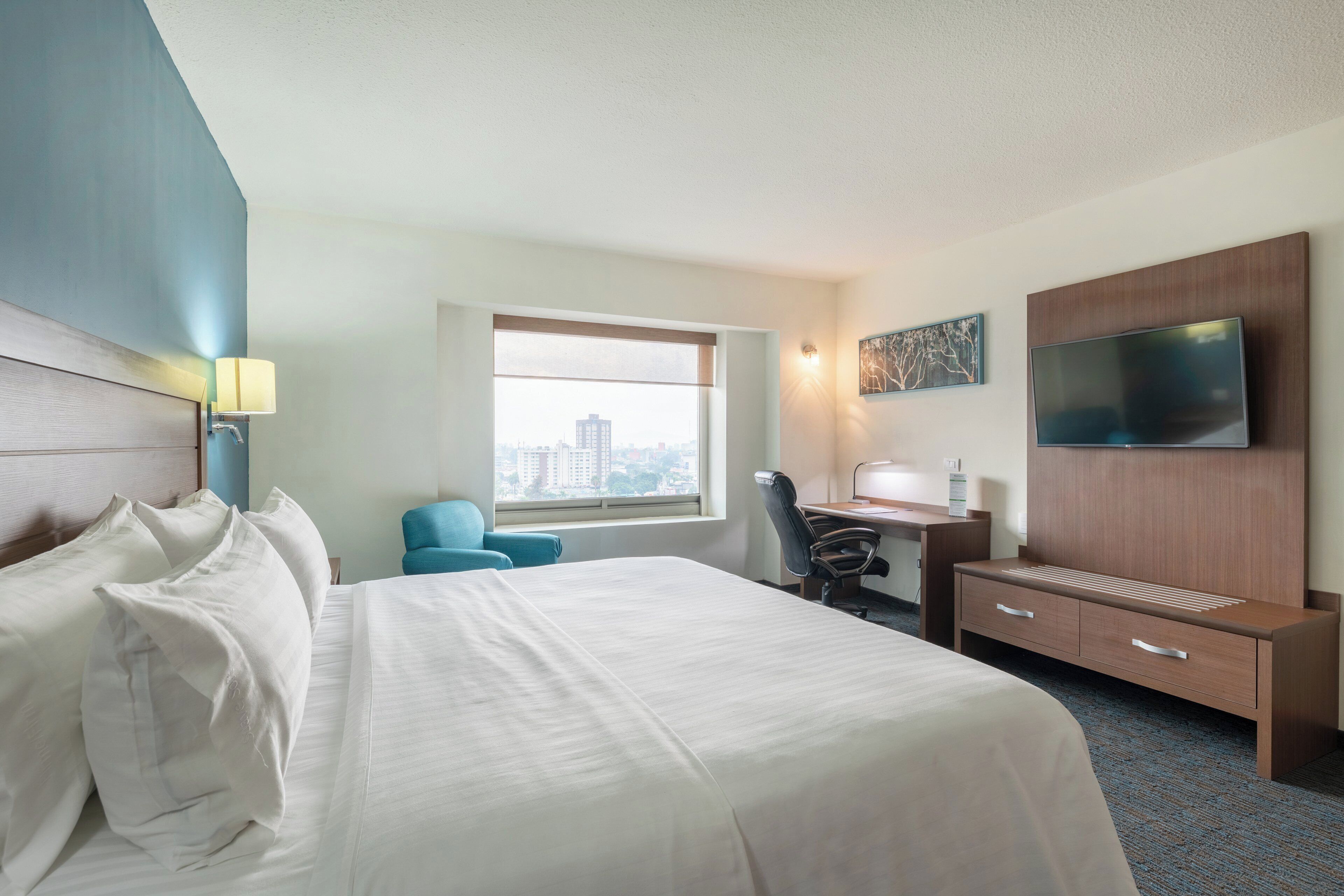 Quarto de hotel com cama, escrivaninha, cadeira, televisão e janela com vista para a cidade.
