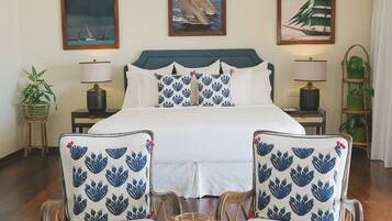 A neatly arranged bedroom with a bed, two chairs with matching cushions, a bedside table with a lamp, and wall art depicting sailboats.