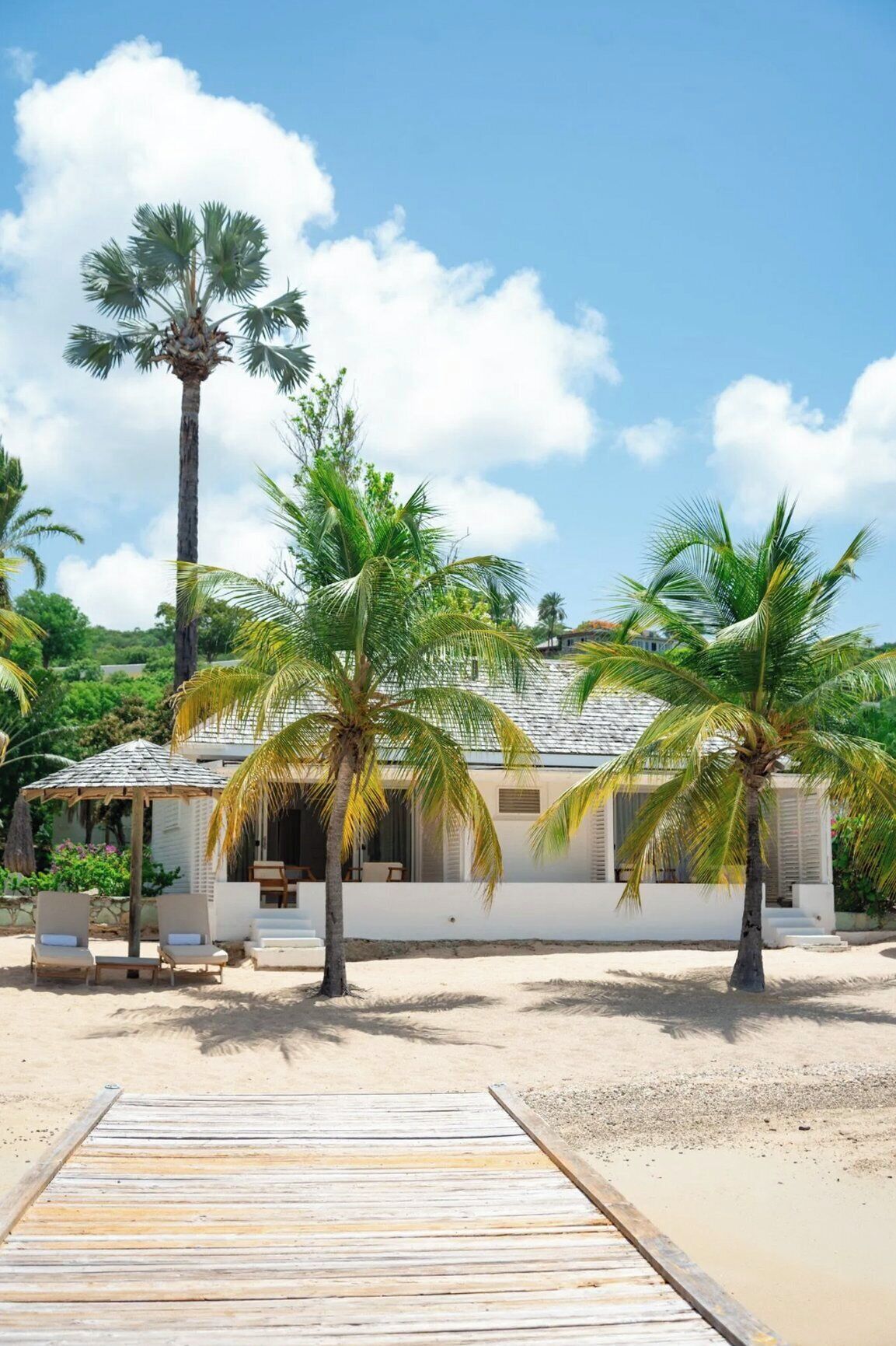 A beachfront property with a wooden deck leading to a white building with a tiled roof, surrounded by palm trees.