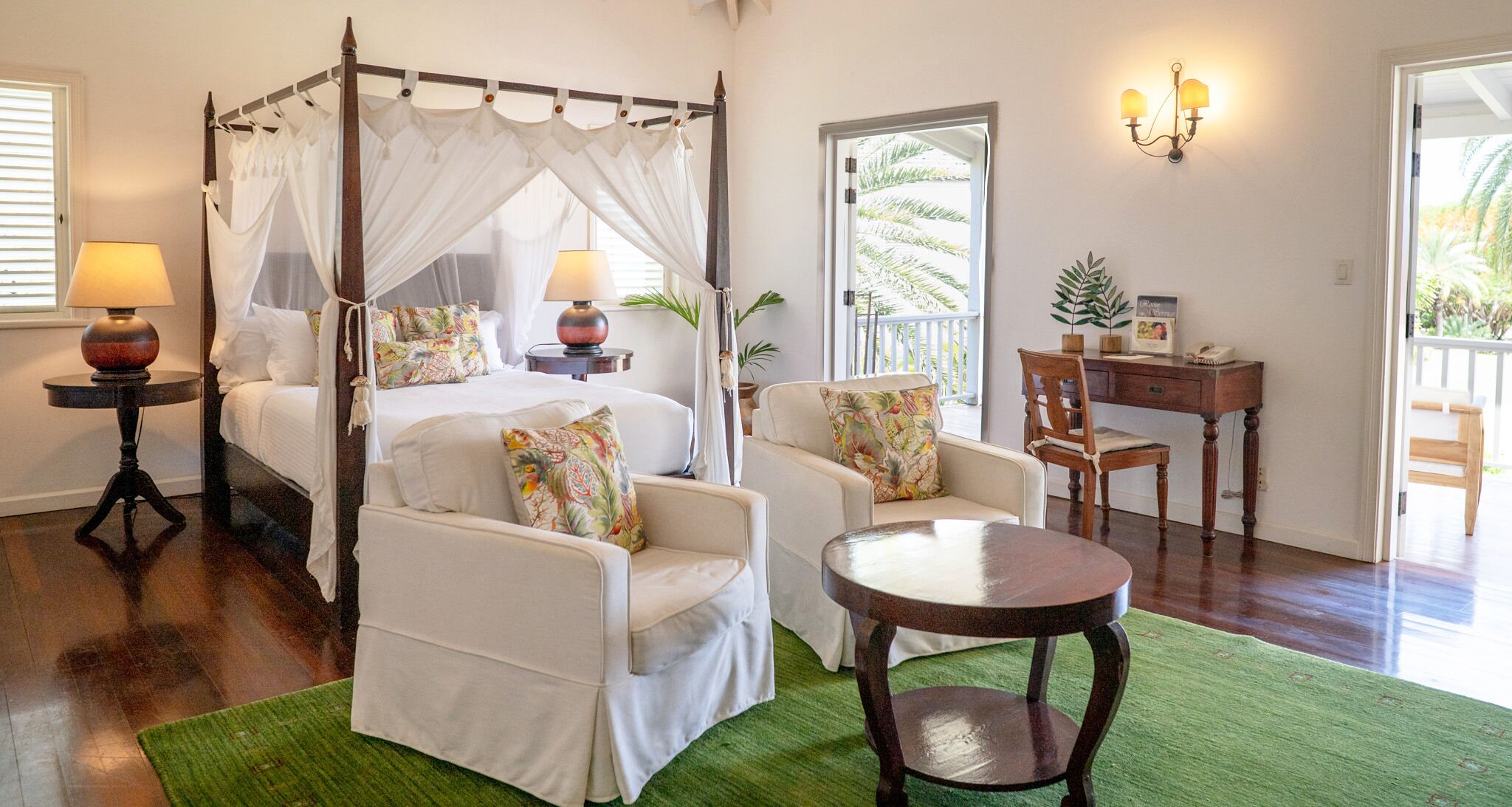 Una cama con dosel y cuatro postes, dos lámparas de noche, una mesita redonda, dos sillones, un escritorio de madera y una alfombra verde en una habitación luminosa con vistas a palmeras.