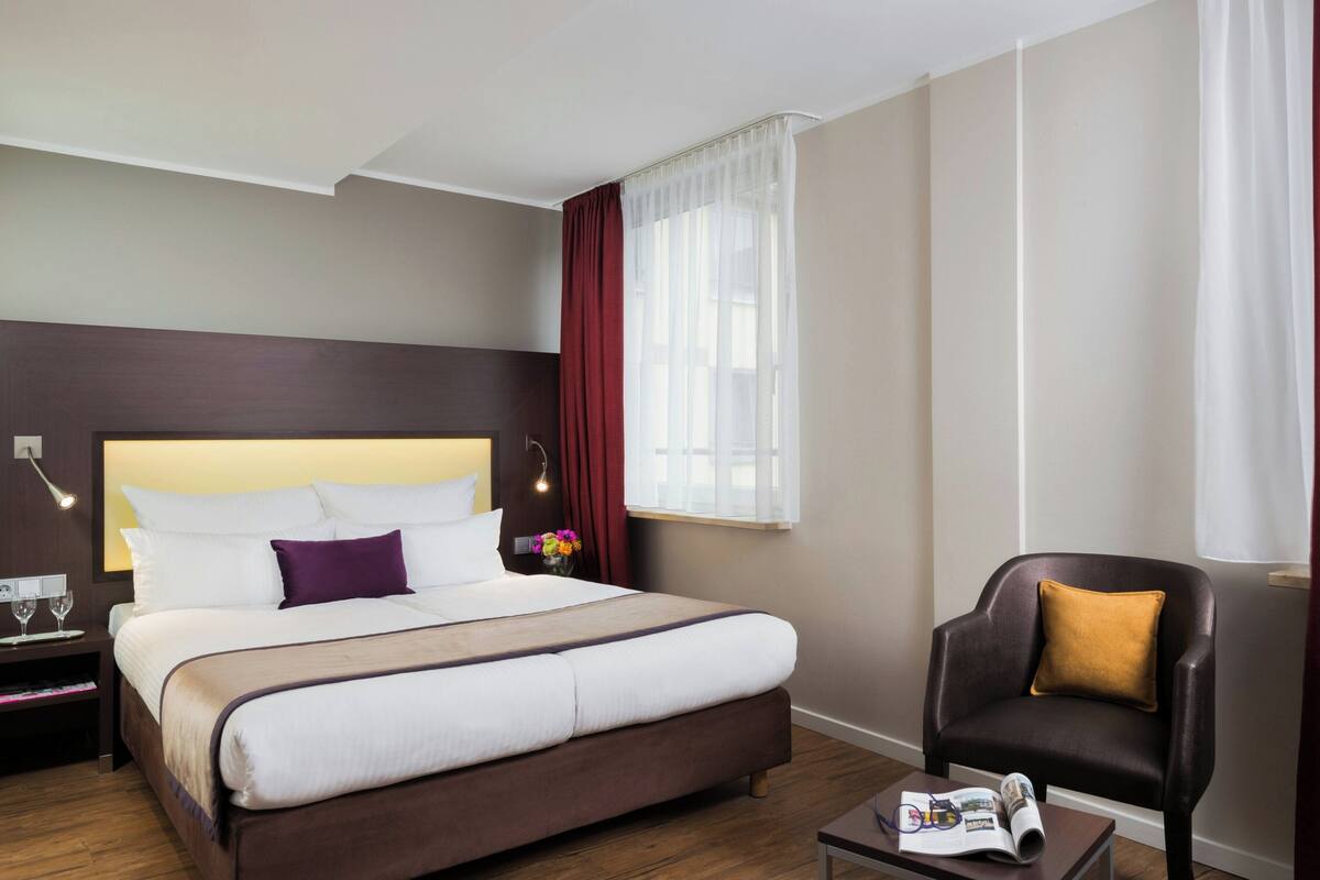 A hotel room with a large bed, a chair, a bedside table with a lamp, and a window with curtains.