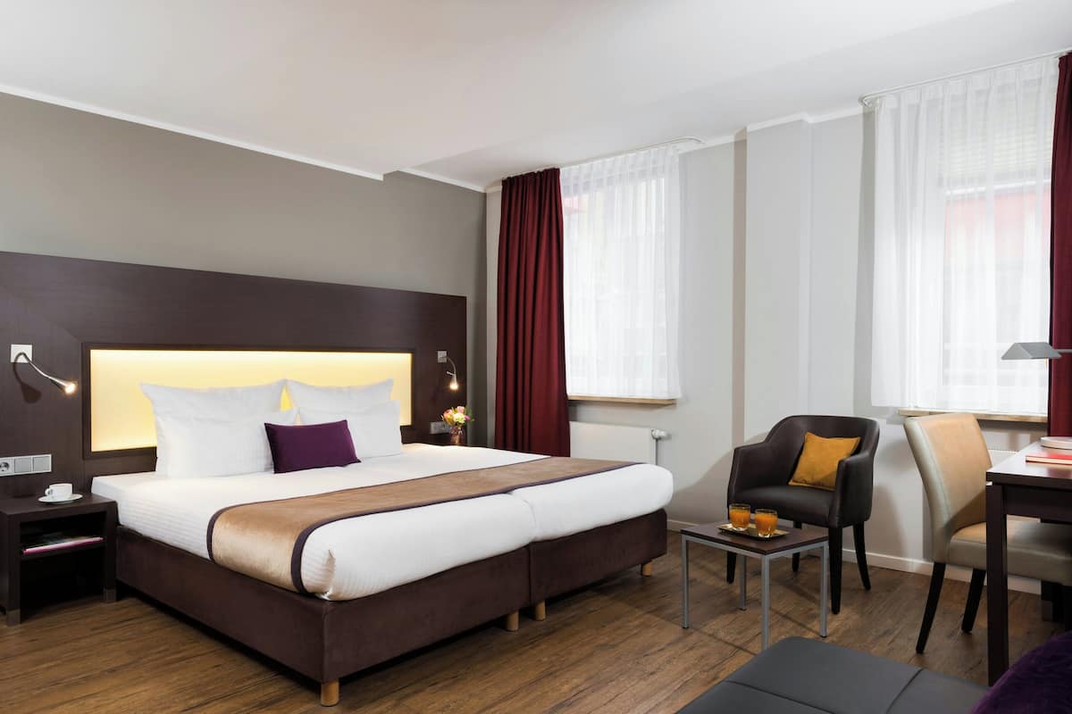 A hotel room with a large bed, a desk, a chair, and a small table with two glasses.