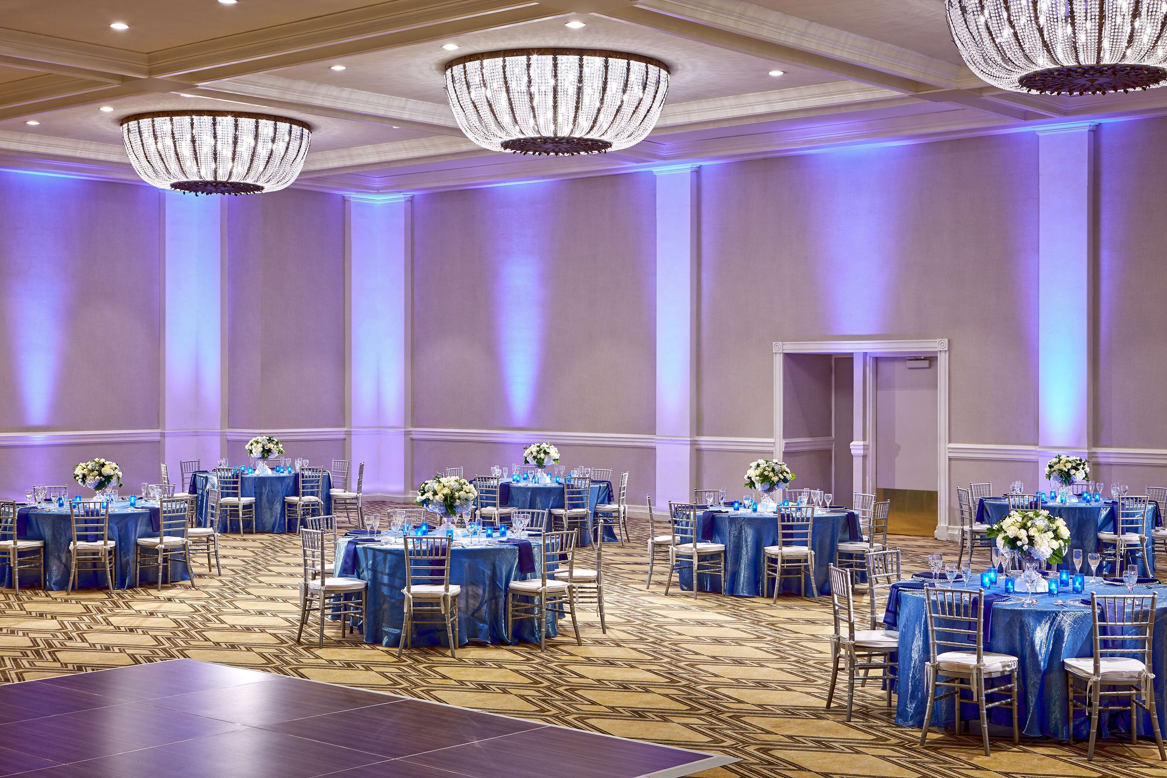 A banquet hall with multiple round tables, each covered with a blue tablecloth and surrounded by chairs.