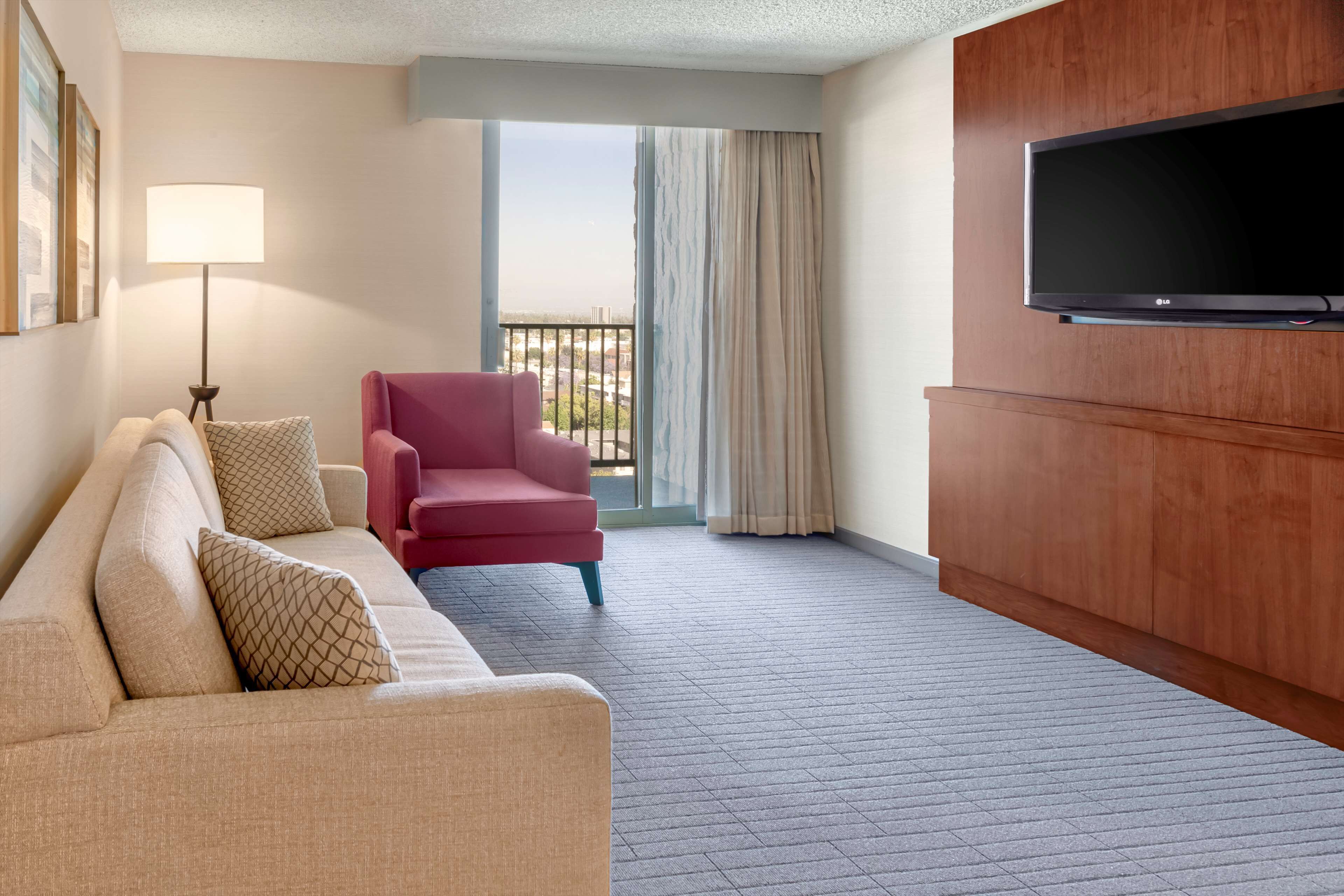 Ein Hotelzimmer mit einem beigen Sofa, einem rosafarbenen Sessel, einem Flachbildfernseher, einer Stehlampe und einem großen Fenster mit Vorhängen.