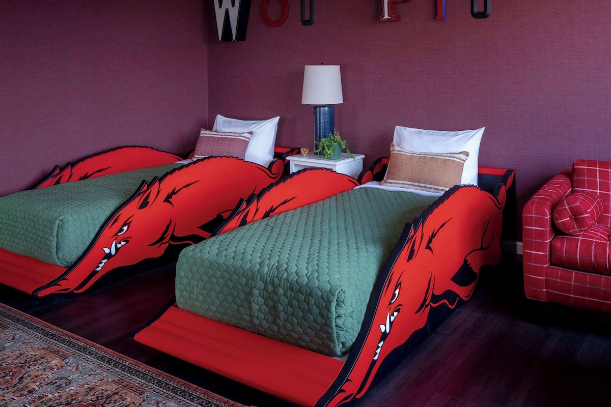 A room with two beds, each with red and green bedding featuring a wolf design. There is a red chair and a small table with a lamp. The wall has letters spelling "Woo Pig".