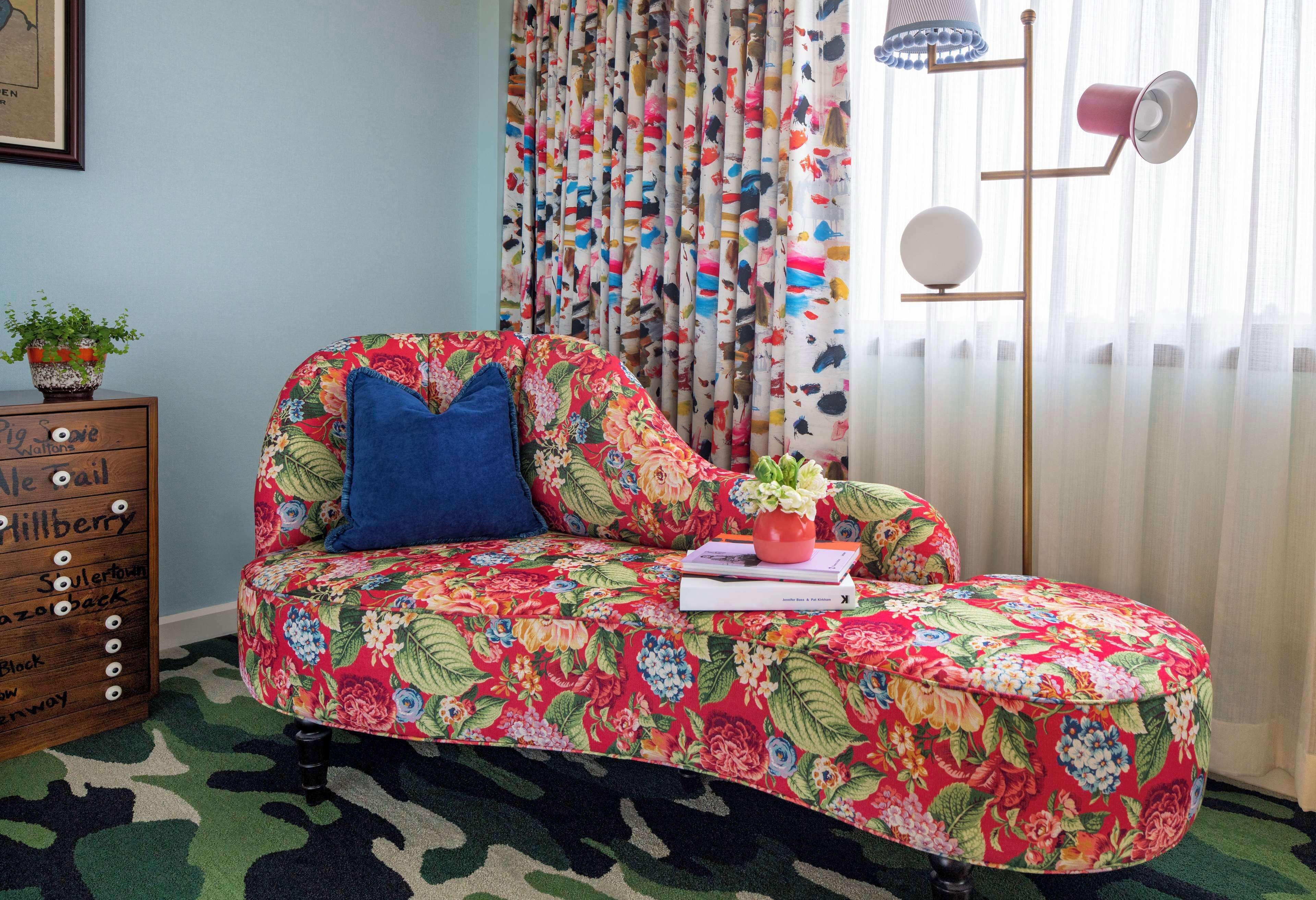 A floral-patterned sofa with a blue pillow and a small potted plant, positioned next to a wooden dresser with drawers labeled with names.