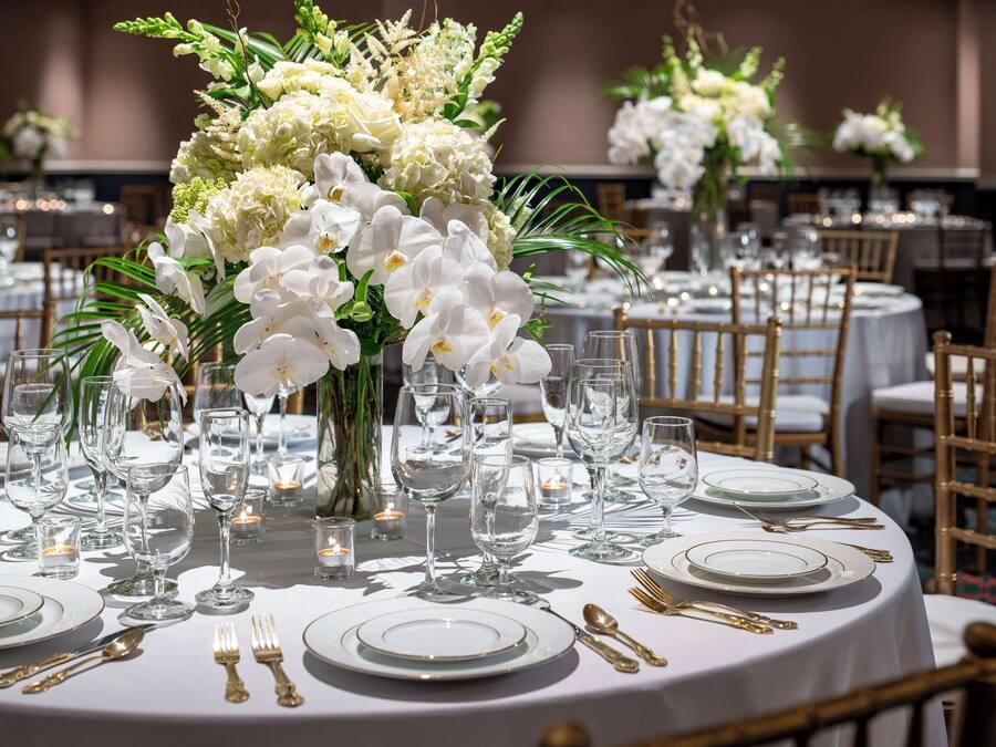 A banquet table set with white plates, gold cutlery, and clear glassware, surrounded by white flowers and candles.
