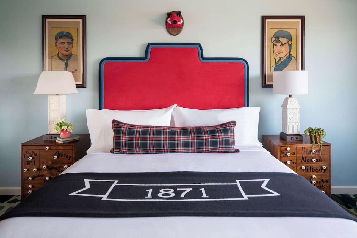 A bed with a red headboard, white pillows, and a plaid throw pillow. There are two wooden nightstands with lamps and decorative items.