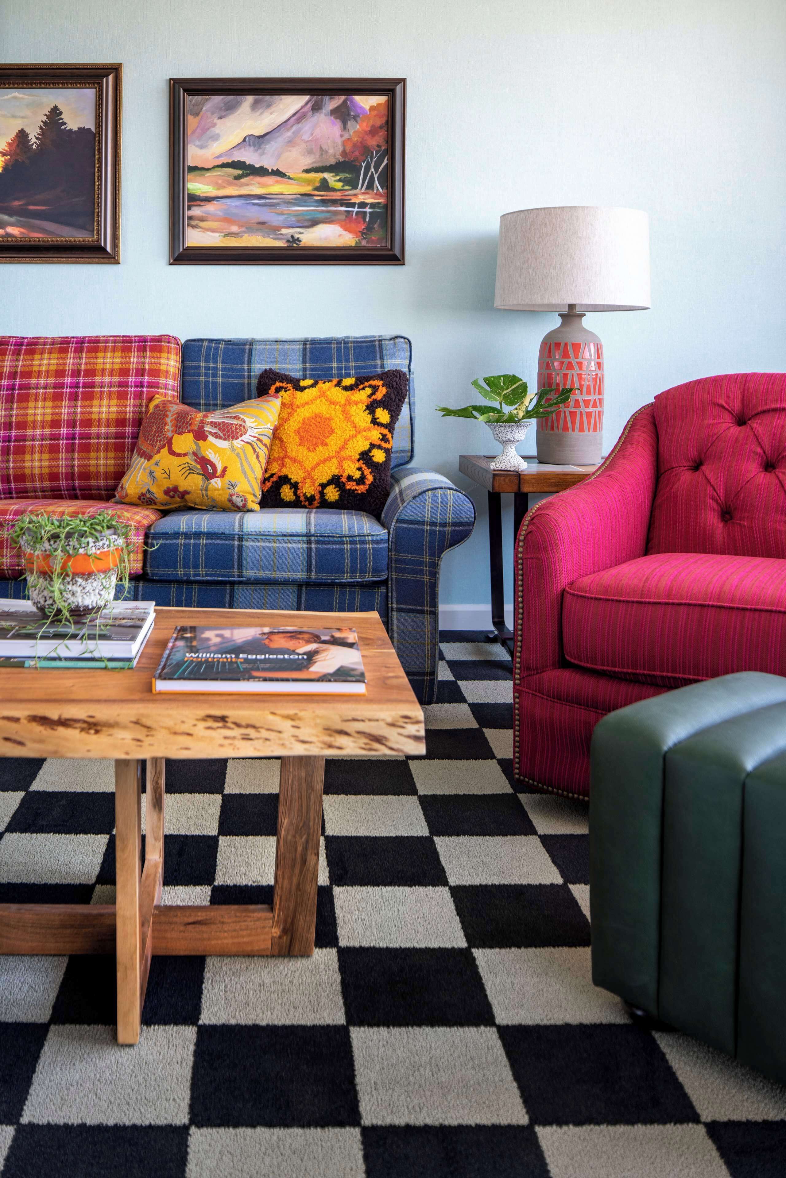 A living room with a checkered floor, a blue sofa with colorful cushions, a red armchair, and a wooden coffee table with books and a plant.