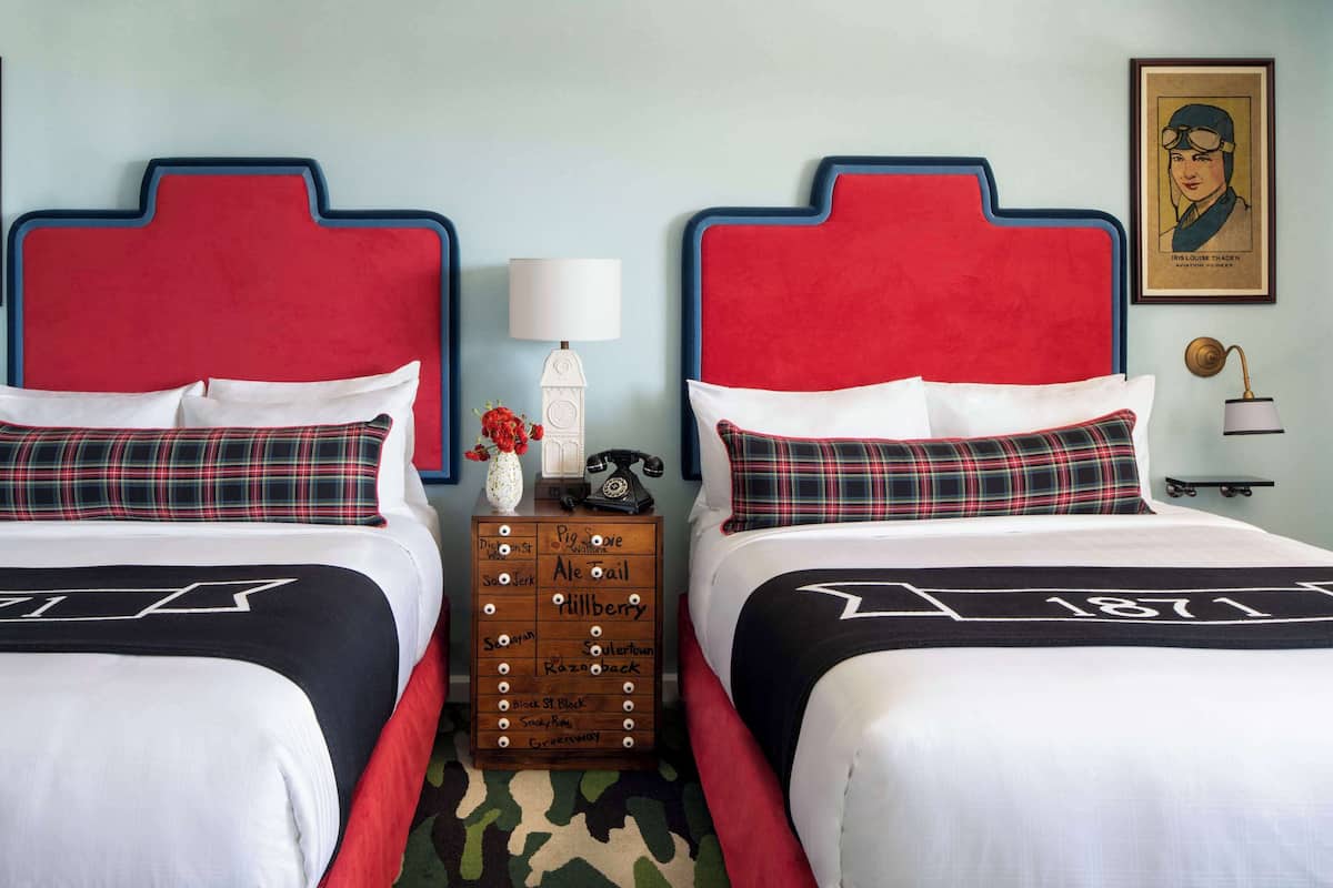 A hotel room with two beds, each with a red headboard and black and white bedding. There is a wooden nightstand with a lamp and a vase of flowers between the beds.