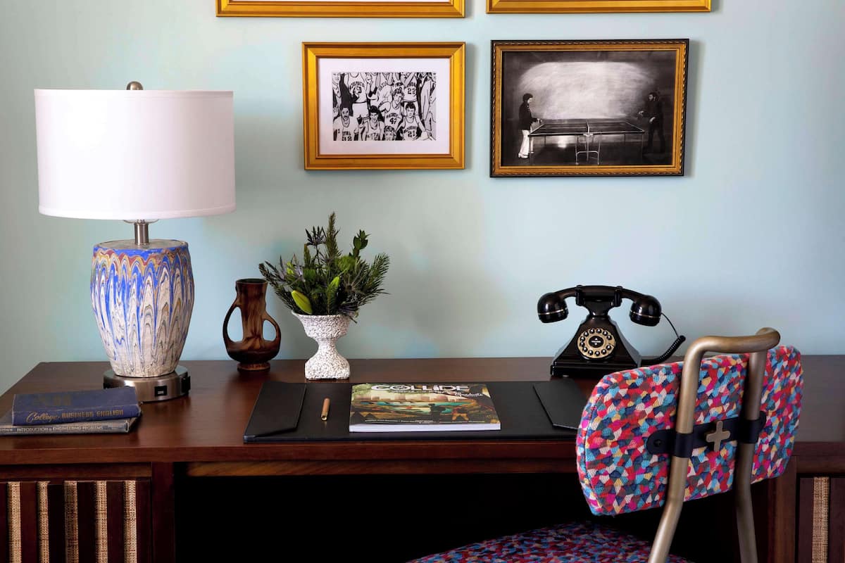 A desk with a lamp, framed artwork, a potted plant, a book, a telephone, and a patterned chair.