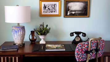 A desk with a lamp, framed artwork, a potted plant, a book, a telephone, and a patterned chair.