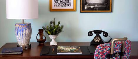 A desk with a lamp, framed artwork, a potted plant, a book, a telephone, and a patterned chair.