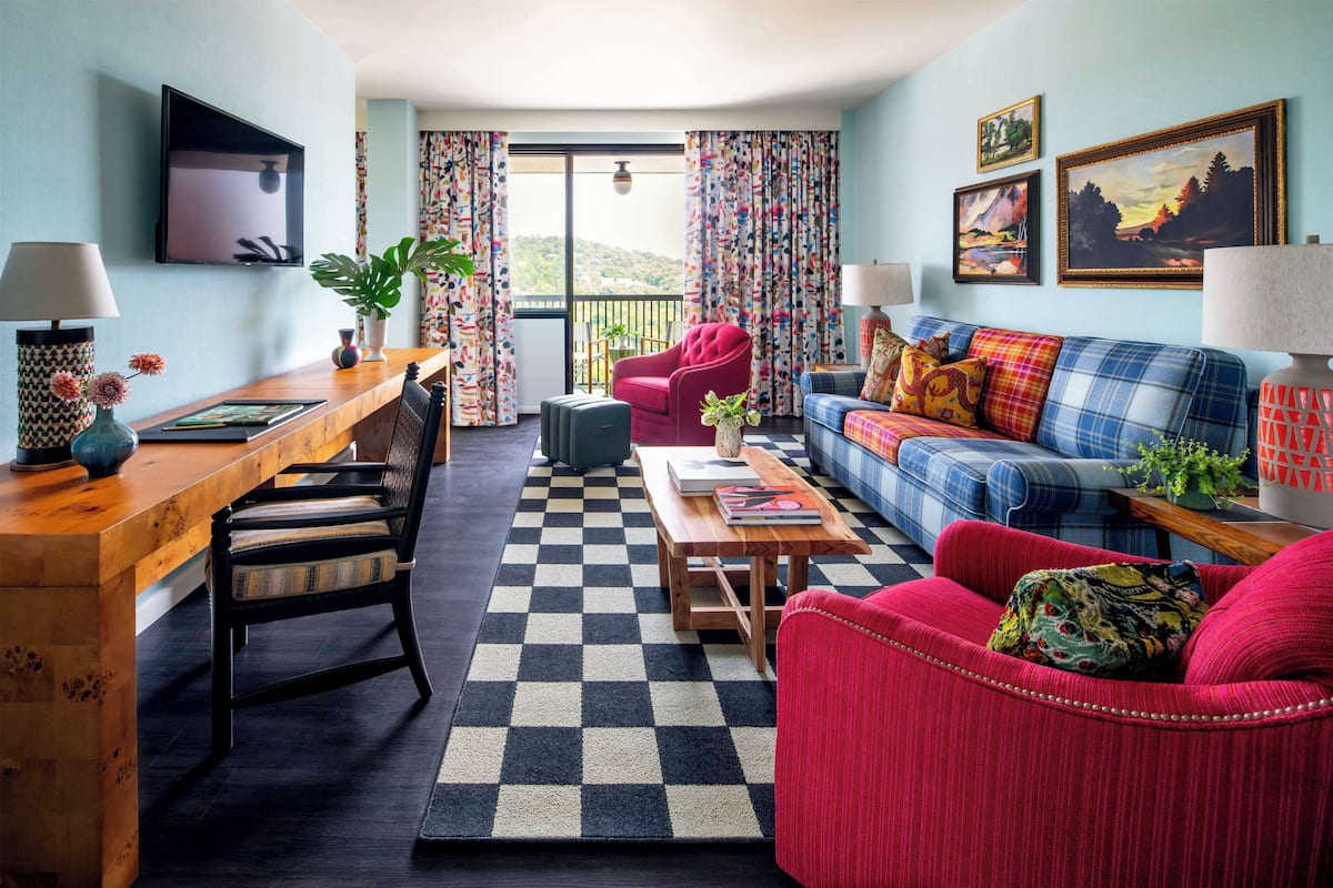 A living room with a checkered rug, a wooden desk, a sofa, and a chair. There are two lamps, a television, and a window with curtains.