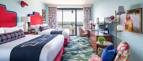 Hotelkamer met twee bedden, een bureau, een stoel, een kleine tafel met een plant en een groot raam met balkon.