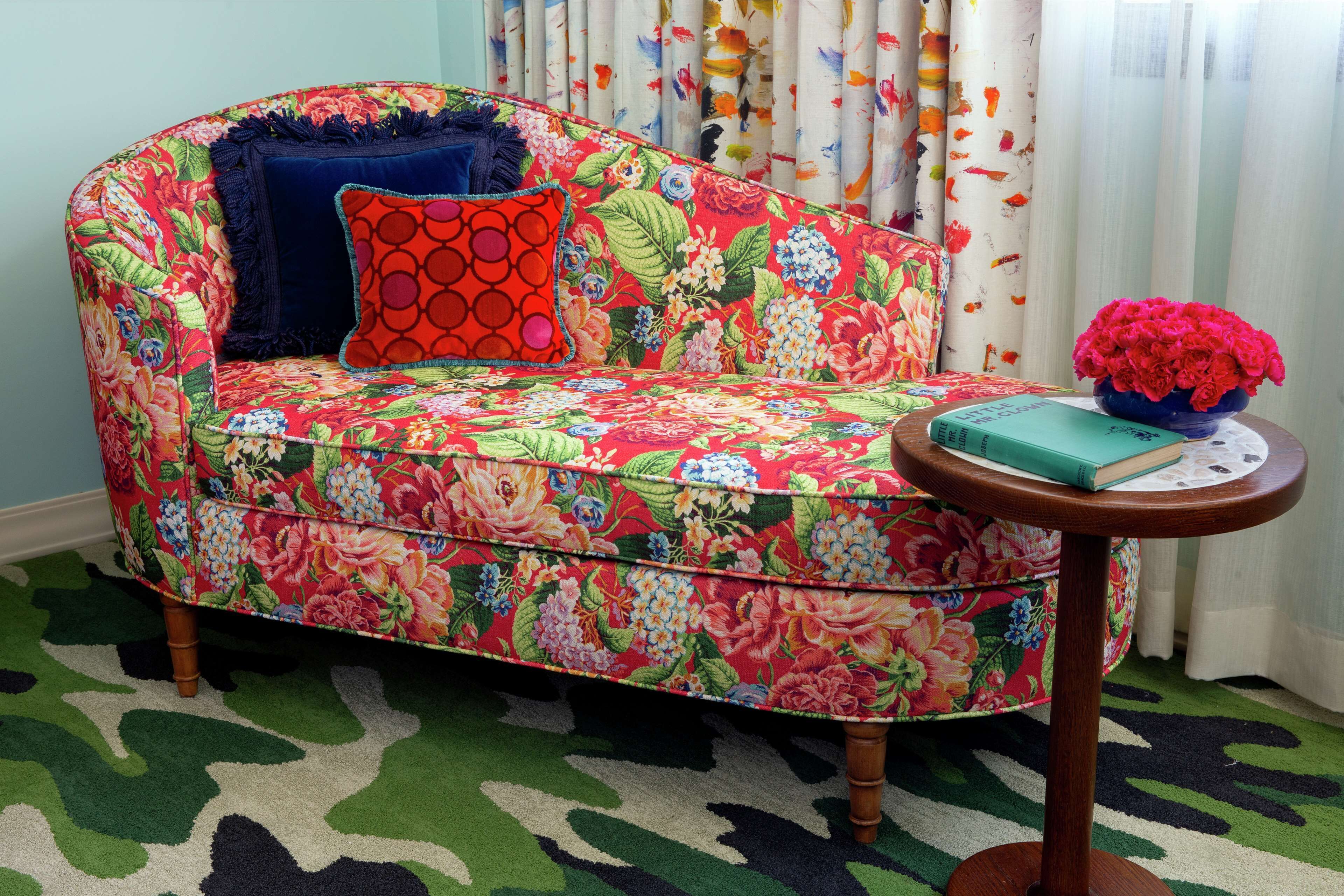A floral-patterned sofa with a blue cushion and a red pillow, a small round wooden table with a book and a vase of flowers, and a patterned rug on the floor.