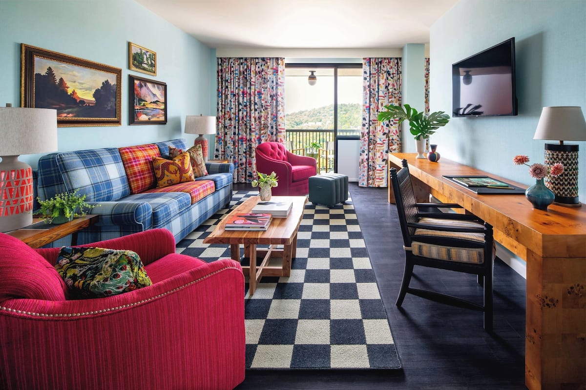 A living room with a sofa, armchair, and a wooden coffee table. There is a TV mounted on the wall and a balcony with curtains.